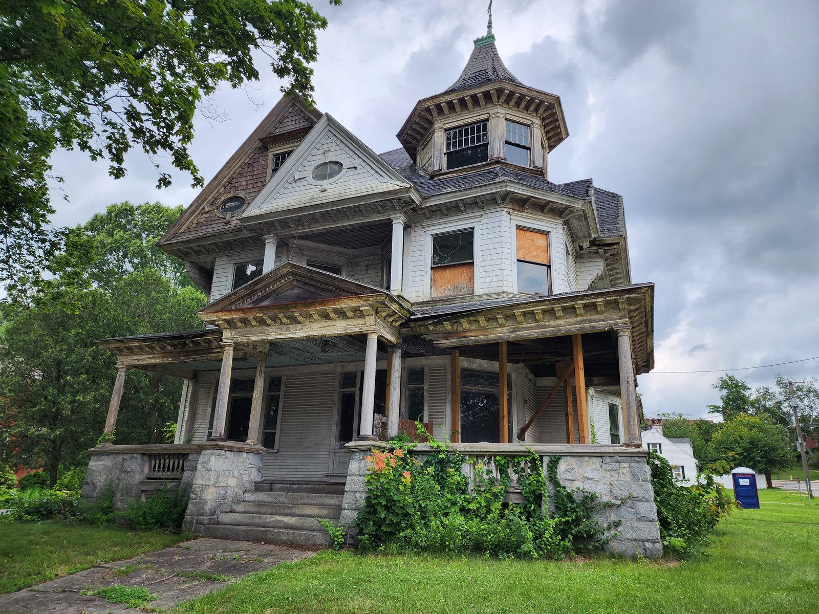 Front elevation before restoration: octagonal corner turret with finial, pedimented porch on classical columns, a window boarded with plywood, granite steps and heavy ivy over the stone foundation