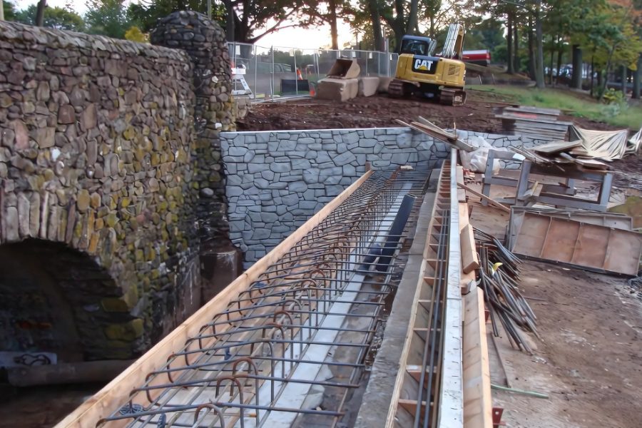 Reinforcing steel set in forms beside the stone arch of the Stanley Quarter pond dam, excavator behind