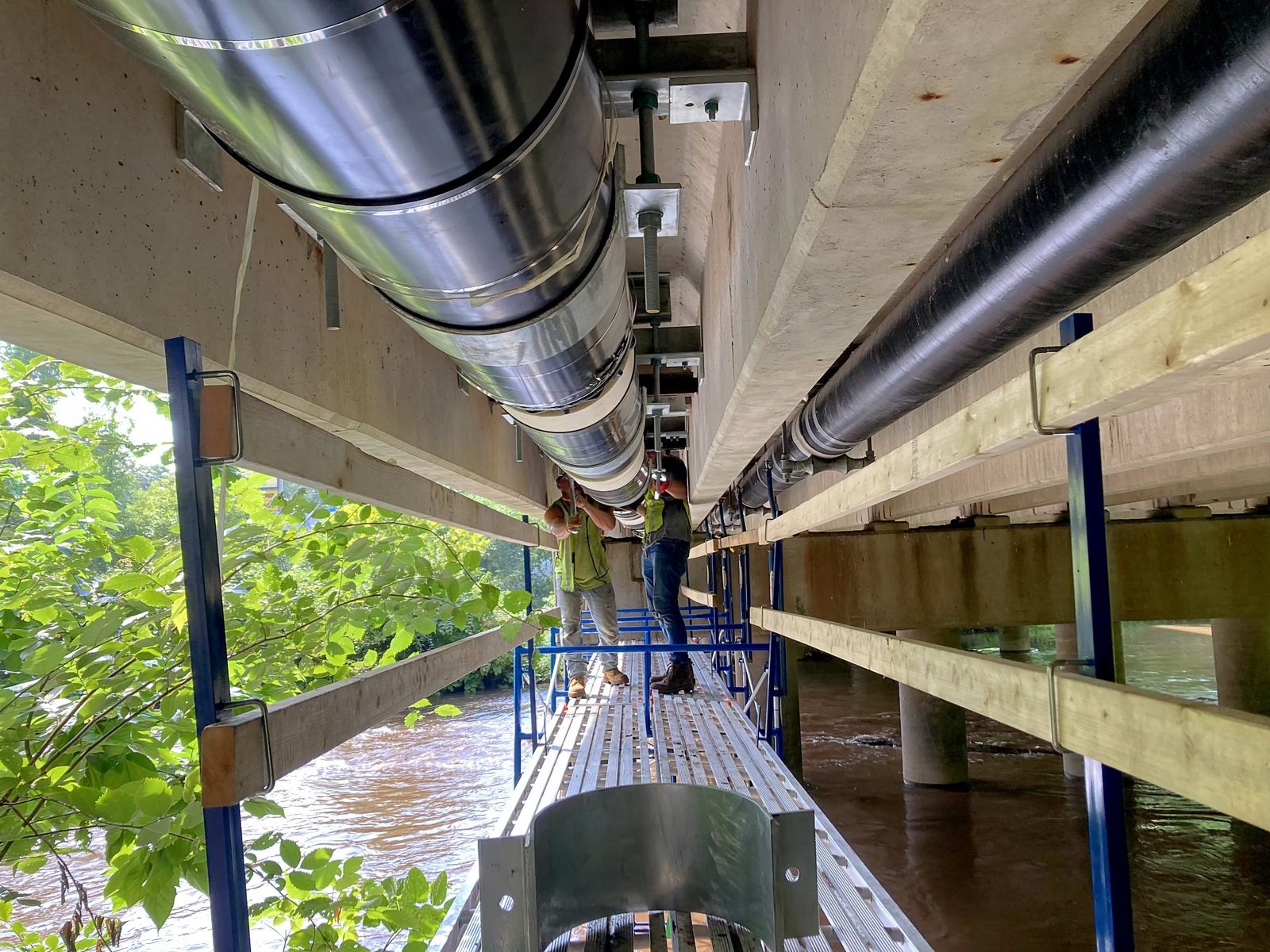 Crew on scaffolding beneath a Wallingford bridge fitting new jacketed insulation and steel hangers to a water main suspended over the river