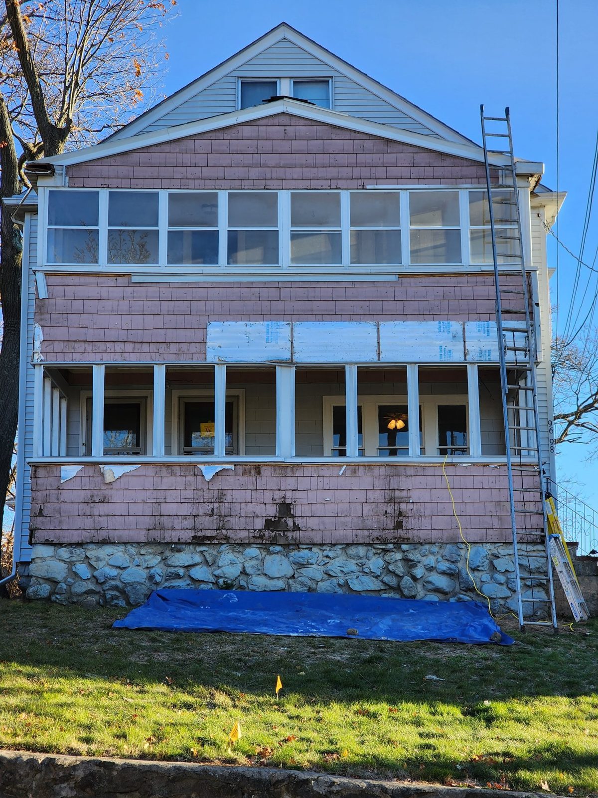 Front of the house with the old shingle siding partly stripped, a ladder against the wall and a tarp on the lawn