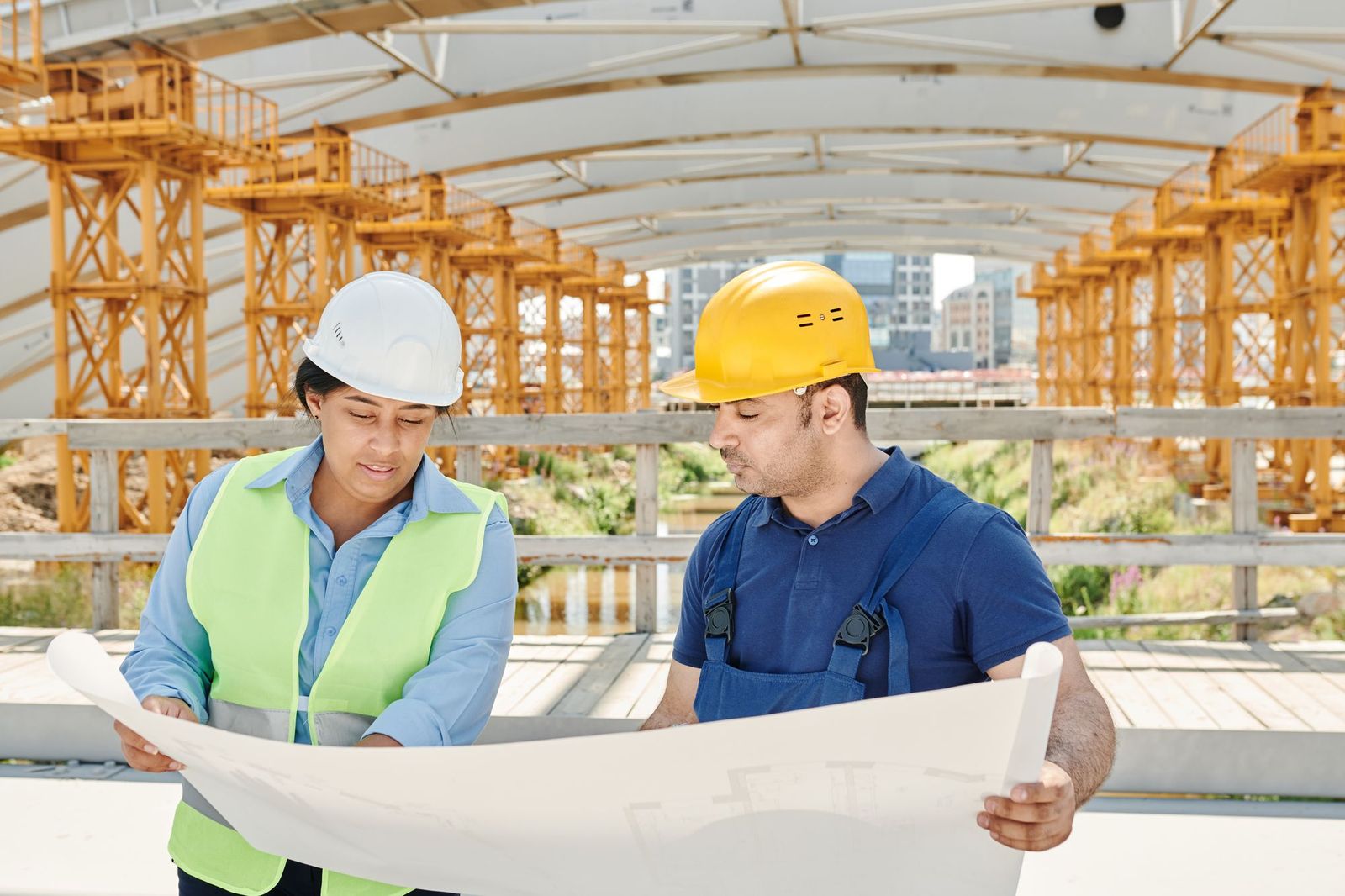 Two construction engineers in hard hats and safety vests unrolling a set of plans on an active jobsite